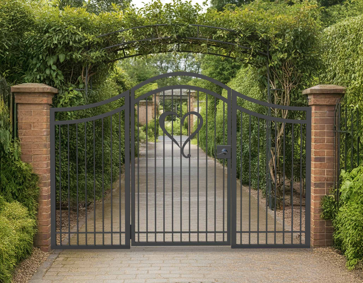 Black powder coated metal pedestrian garden gate with side panels featuring central heart motif and arched top frame installed between brick pillars