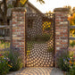 Laser cut metal garden gate with a cobblestone spiral vortex pattern in matte sepia brown (RAL 8014), installed between reclaimed brick pillars at a rustic farmhouse entrance.