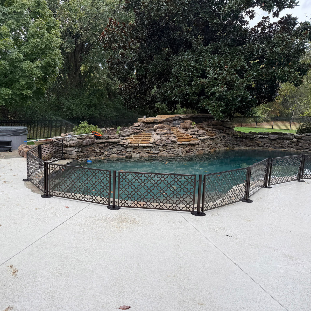 No drill freestanding dog pool fence installed on a concrete pool deck without anchors, panels angled around a rock waterfall pool