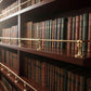 Solid unlacquered brass gallery rail rods mounted on multi-shelf mahogany wood library bookcase holding vintage leather-bound books.
